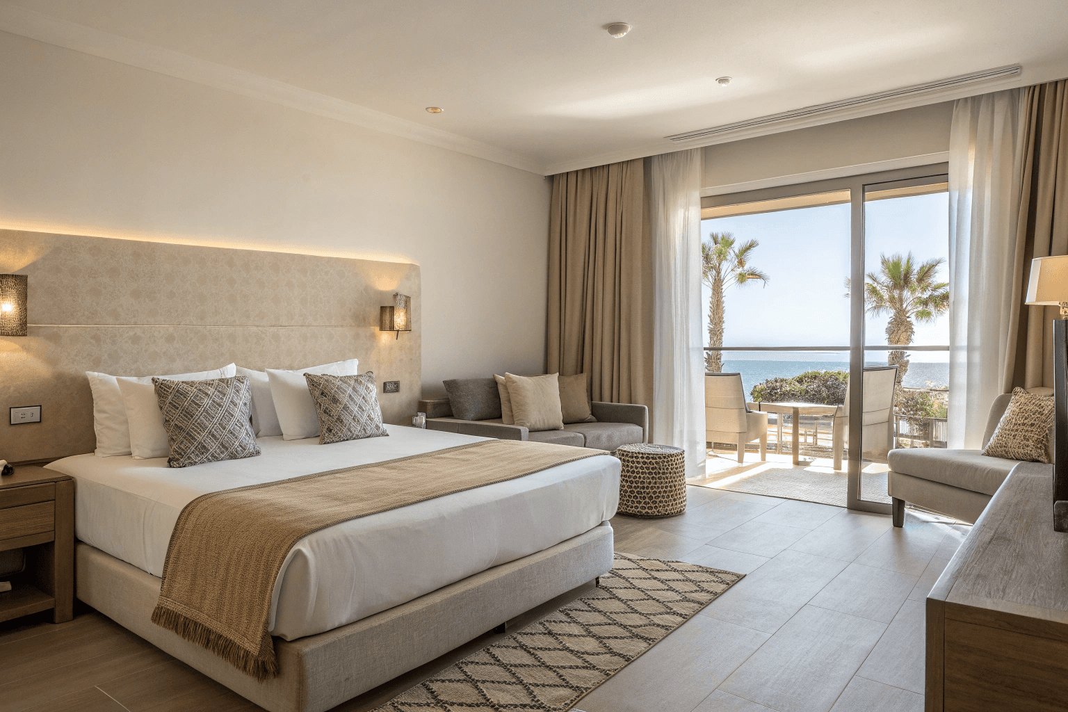 sleeping and living areas, king bed with layered neutral linens, elegant reading lights, and modern sofa with textured cushions